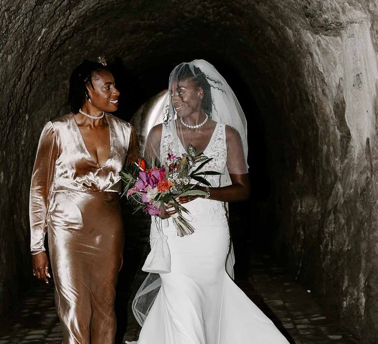 Bride in v neck sleeveless lace Pronovias wedding dress, cathedral length veil and silver sparkly bridal jewellery holding colourful tropical African bridal bouquet walking with mother of the bride in long sleeve golden silk wrap around dress and black fascinator through tunnel at Tunnels Beaches 