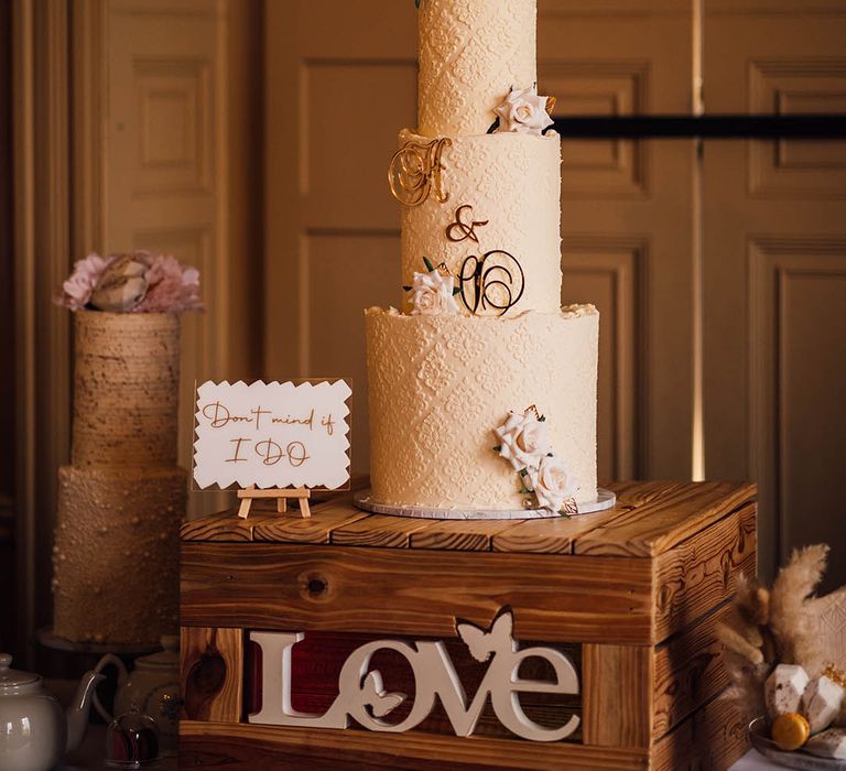 Three tier buttercream frosted wedding cake with delicate pattern, iced pink roses and personalised gold lettering 