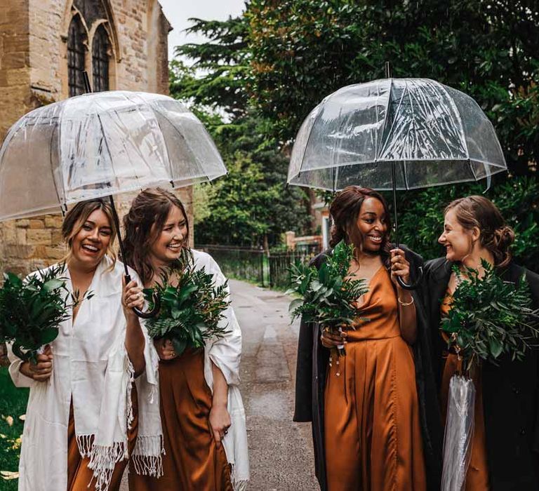 Bridesmaids in strappy sleeveless front slit burnt orange bridesmaid dresses holding foliage bouquets and shawls standing underneath umbrellas outside of church wedding