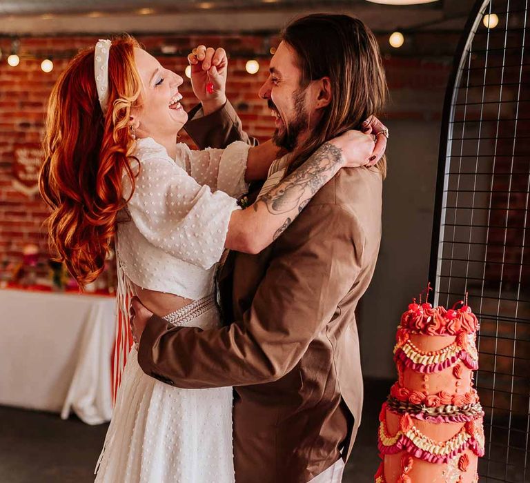 Groom in brown grooms blazer feeding bride maraschino cherry in cut out back detail short sleeve wedding dress standing by retro wedding cake at Northern Monk Refectory Leeds