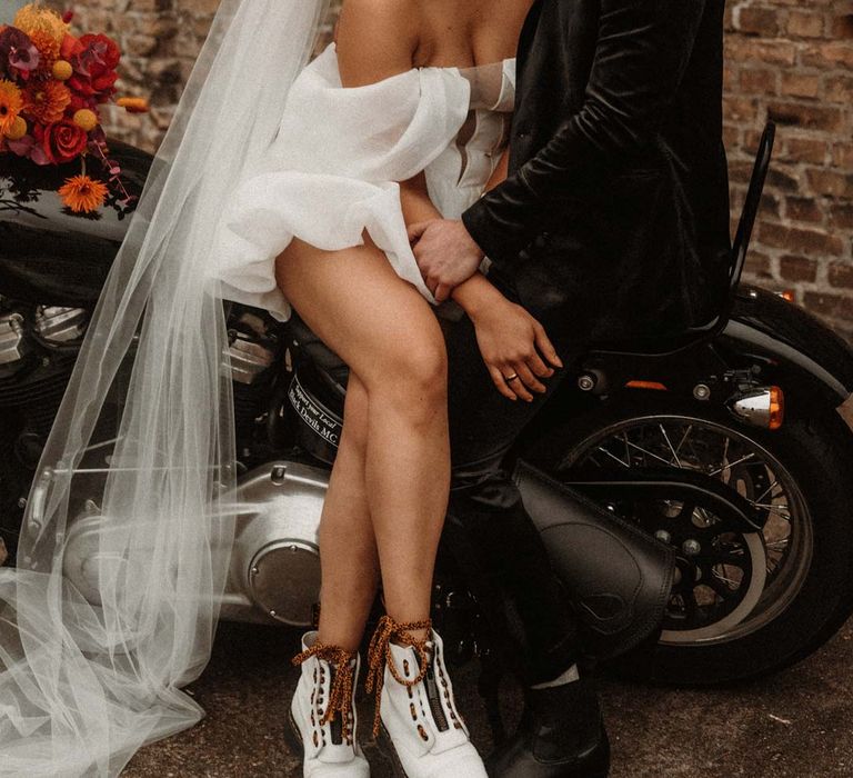 Groom in wet look black grooms suit with leopard print shirt and pink and orange boutonniere standing with bride in off shoulder tulle short wedding dress with church length veil and white platform Dr Martens wedding shoes