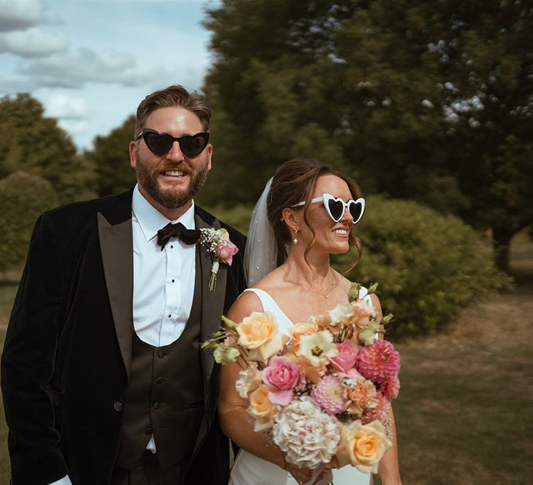 Bride in white wedding satin slip dress with colourful bouquet and retro white sunglasses with groom in black tuxedo with sunglasses