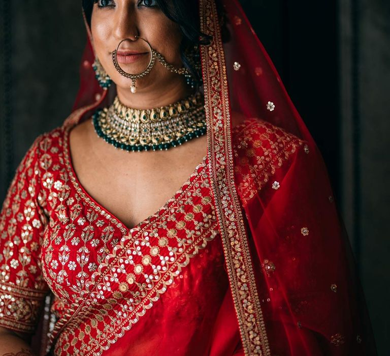 Bride in red and gold lehenga, gold and forest green bridal jewellery, and gold nath 