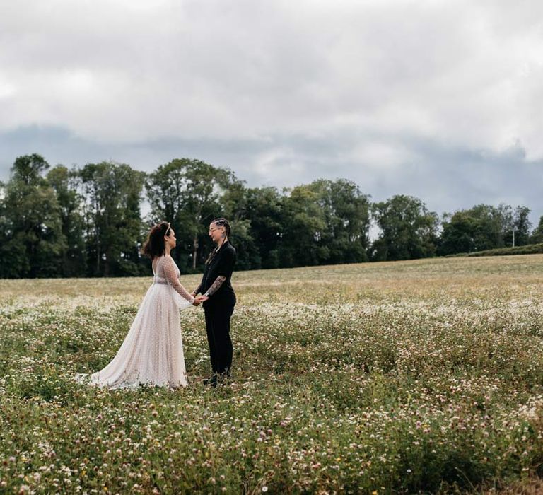 Bride in long sleeve v-neck sheer polka dot overlay sparkly wedding dress and gold celestial bridal headband holding hands with bride in silver chains, glasses, black shirt and black trousers at Viking themed wedding