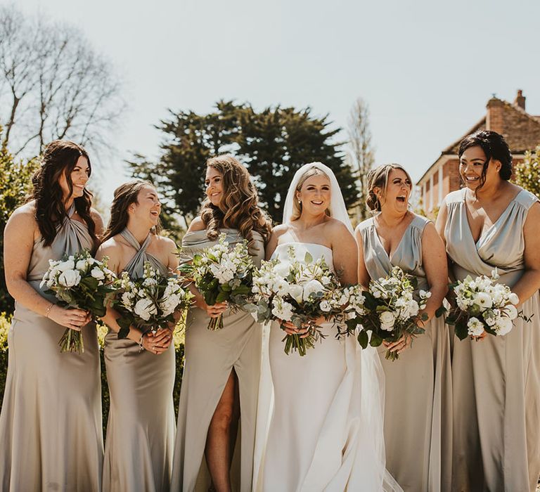 Bridal party in golden satin multiway mismatched style bridesmaid dresses with the bride holding matching white bouquets 