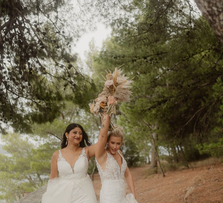 two bride in v front wedding dresses holding dried flower bouquets in the air 