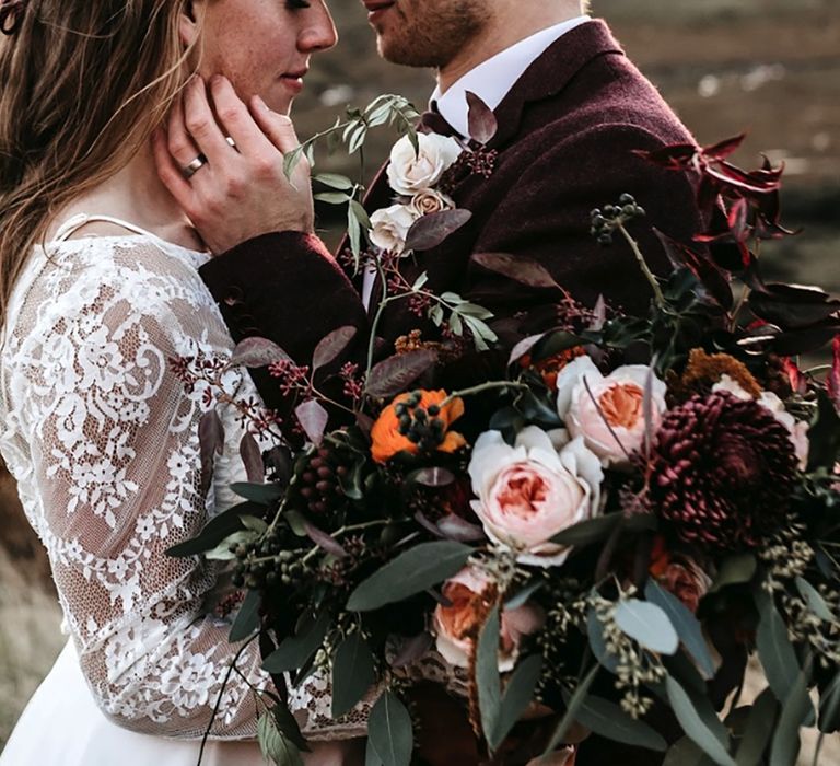 Dark deep red autumn wedding leaves in bouquet held by bride in lace wedding dress with groom in burgundy suit 