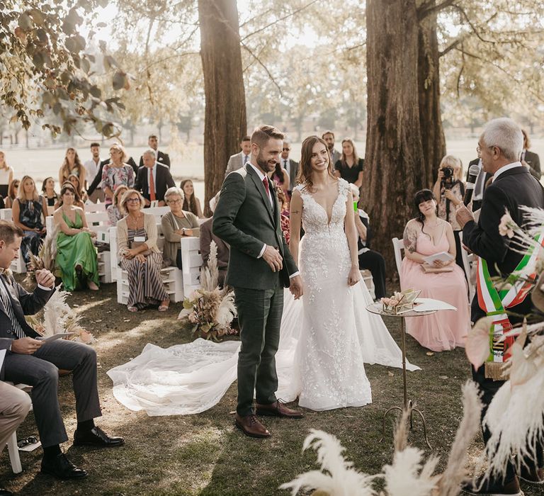 bride in an appliqué wedding dress holding hands with her groom in a dark green suit at their outdoor tenuta il cigno wedding with pampas grass floral arrangements 