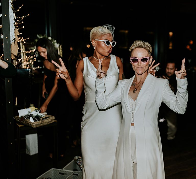 Bride wearing satin wedding dress with bride in blazer floor length jacket over two piece outfit with sunglasses for wedding reception 