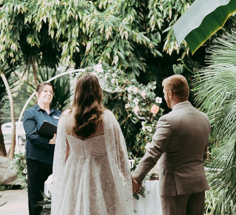 Bride in embroidered wedding dress and cape standing hand in hand with groom in grey suit at wedding 