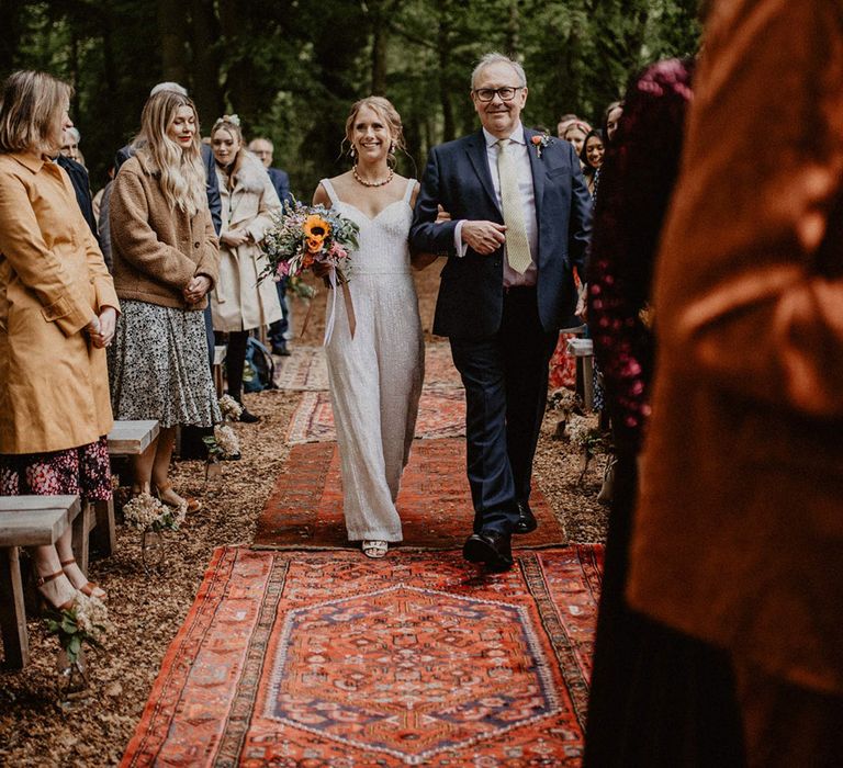 The bride in a jumpsuit walks down the aisle with the father of the bride 