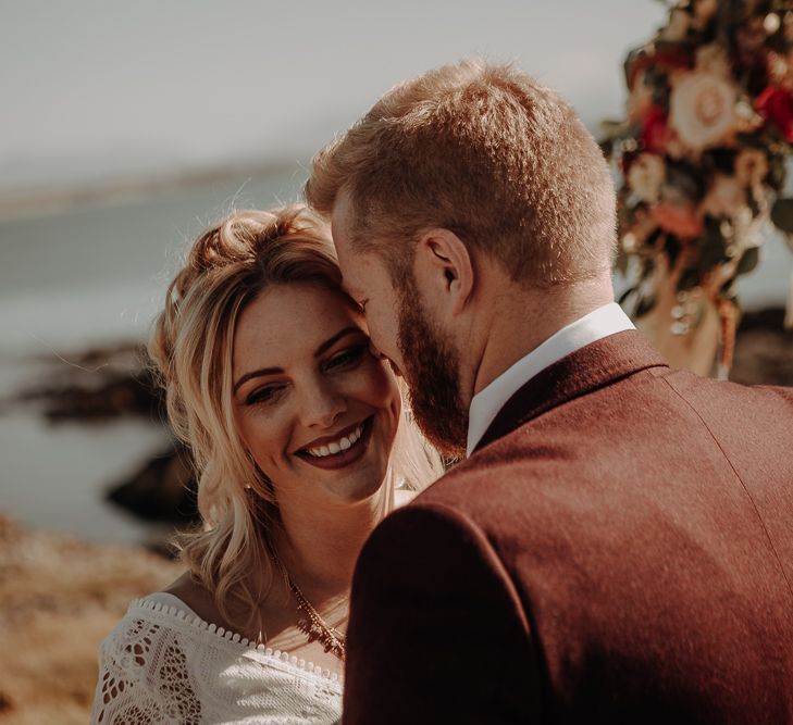 Bride and groom in burgundy suit pose on Anglesey Island with boho decor in the background