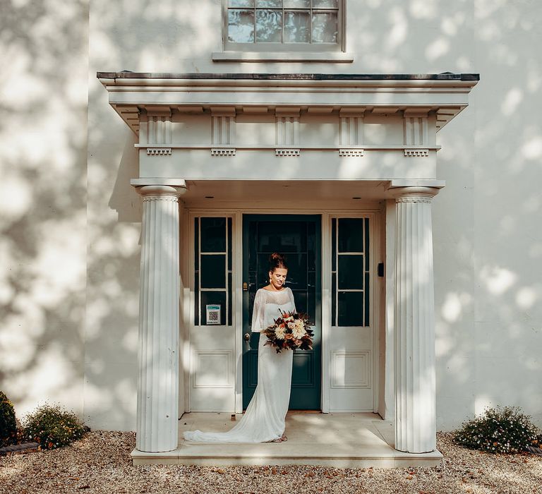 Bride with brown hair and red lipstick stands with bouquet in front of white house