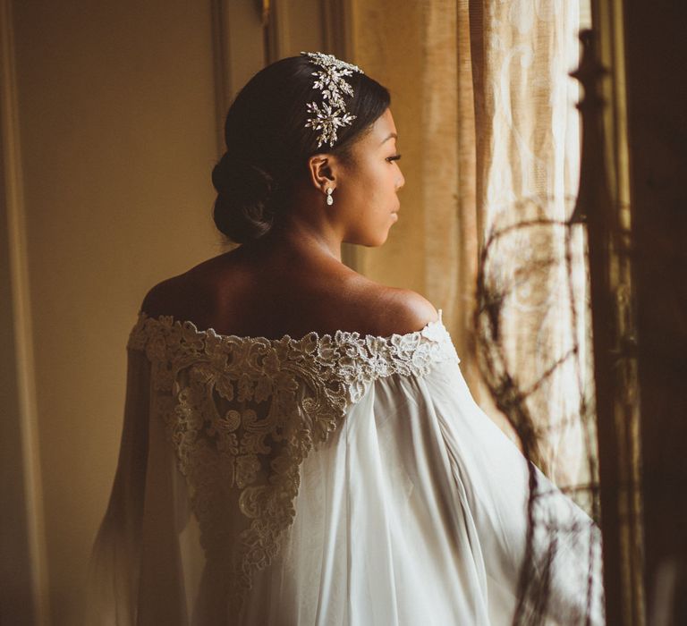 A bride with a low bun in her hair looks out of the window. Her wedding dress has a sheer cape.