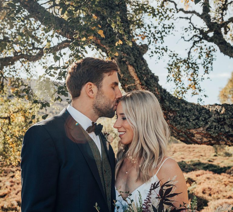 Groom kisses bride on the forehead whilst she holds Autumnal coloured bouquet 