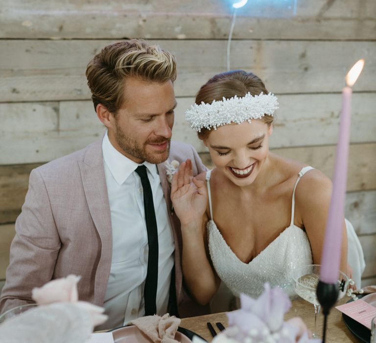 bride in a pearl covered headband wearing a thin strapped jumpsuit sitting at a table with neon sign backdrop