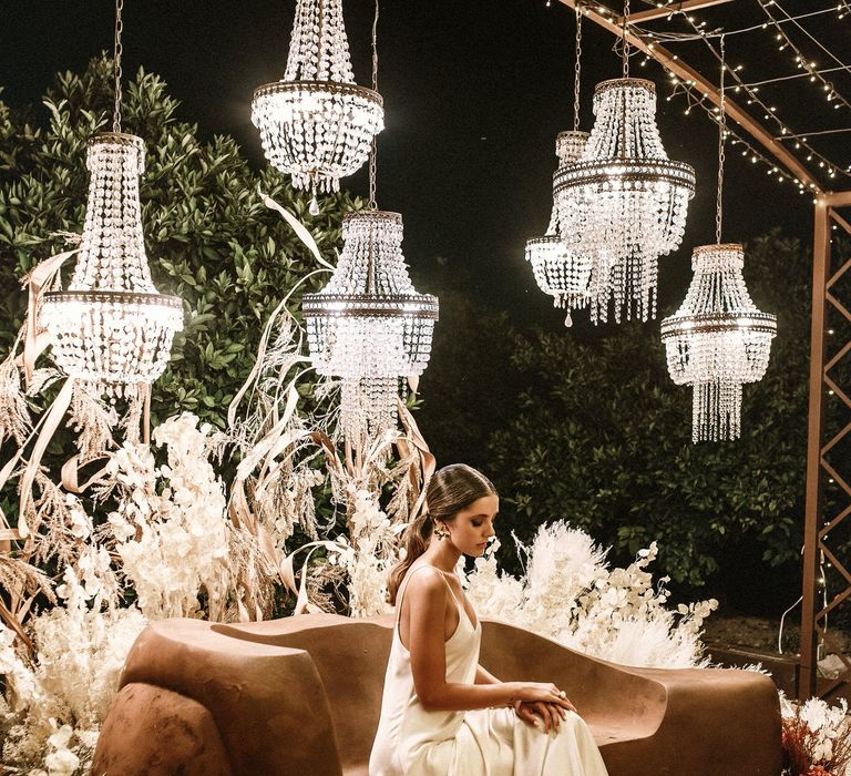 Bride in a satin slip wedding dress sitting under a canopy of lights and chandeliers with a floor full of wicker rugs 