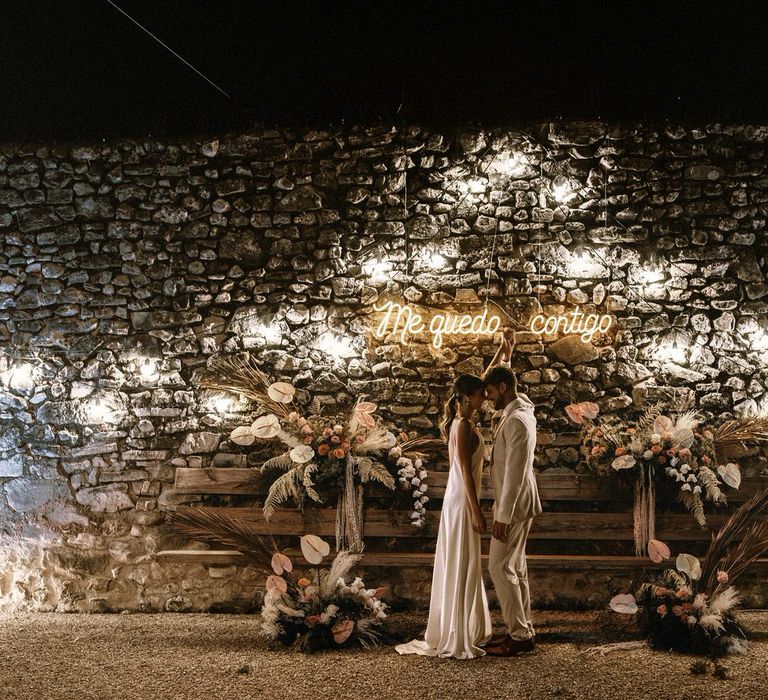 Outdoor seating area with the bride and groom standing in front of an exposed brick wall decorated with a neon sign, festoon lights and boho wedding flowers