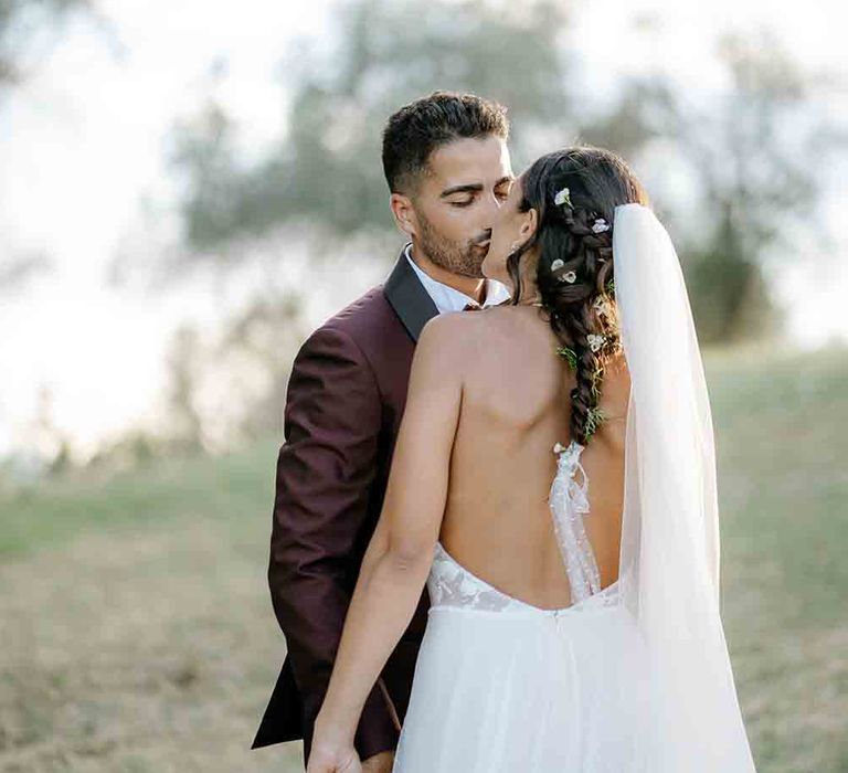 Bride clutches large bouquet wearing backless wedding dress with groom in maroon suit