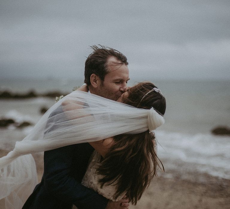 Bride & groom embrace with the sea in the background and brides veil blows around them