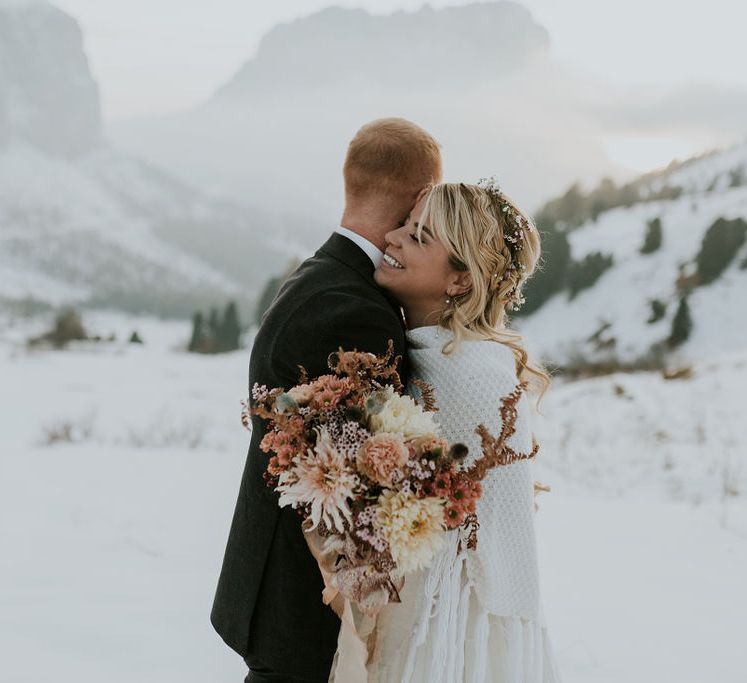 Bride and groom embracing on the snowy Dolomite mountains wearing a shawl and delicate flower crown and holding an orange autumn wedding bouquet 