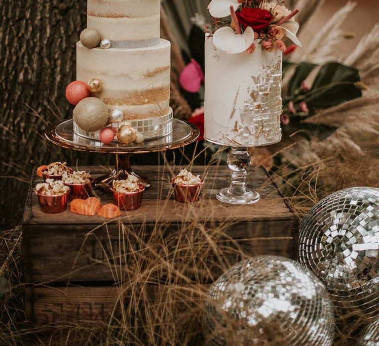 Three tiered naked wedding cake with white icing, silver ribbon and ball decorations on wooden table next to single tiered white column wedding cake with silver detailing and floral decor at festival themed wedding