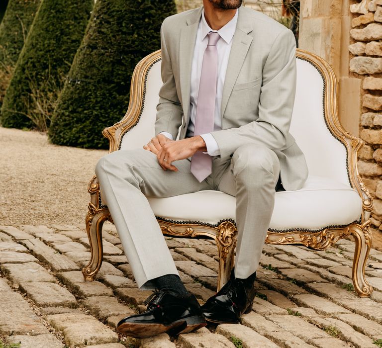 Groom sits on white and gold chair whilst wearing pale suit and pale pink tie 