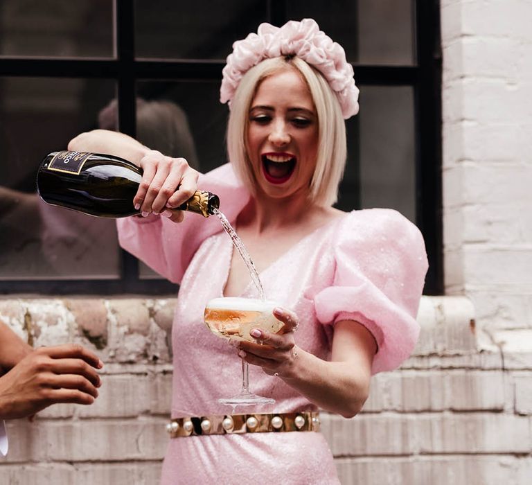 Bride in a custom-made pink jumpsuit and appliqué headband pouring champagne