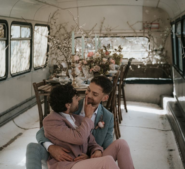 Grooms sit beside wooden table in rustic setting with pastel floral decoration and brass candle holders  