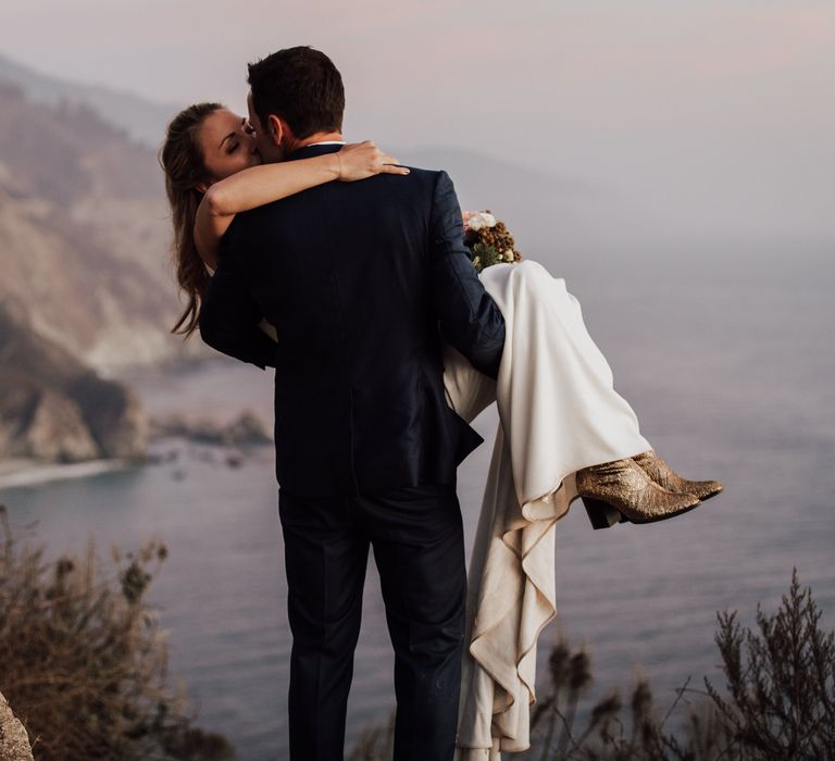 Groom carries bride along the cliffside as the ocean can be seen in the background