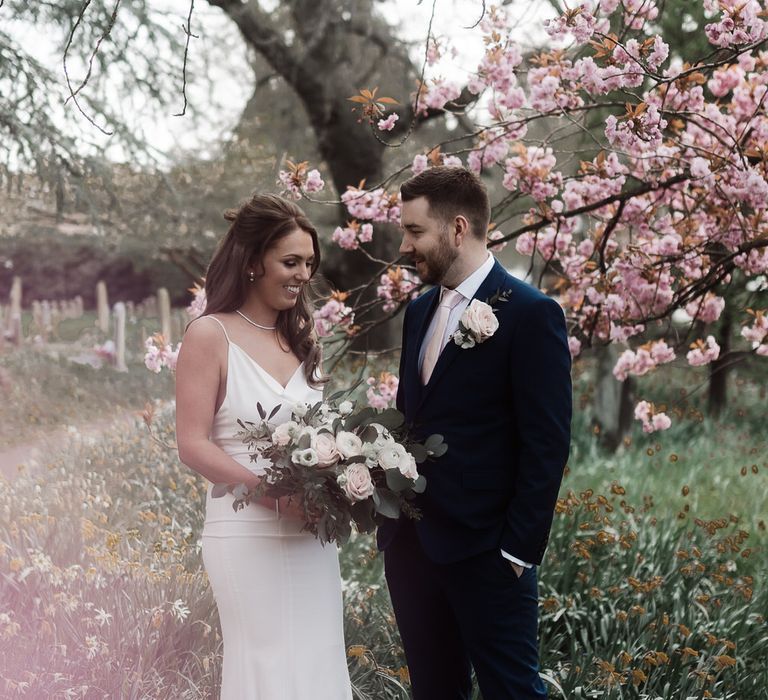 Bride in a white slip wedding dress and groom in a navy suit stand in church grounds surrounded by blossom