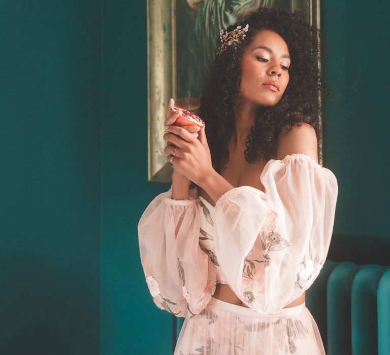 Black bride with long curly hair in blush pink bridal separates holding a pomegranate