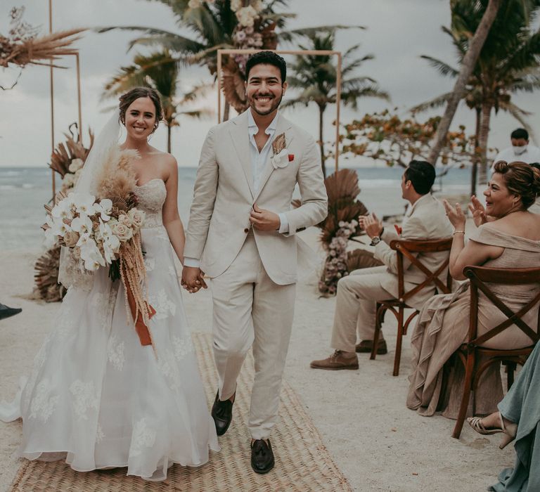 Bride & groom walk down the aisle after wedding ceremony on the beach as aisle is lined with dried florals