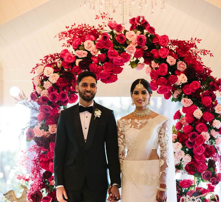 Bride & groom stand in front of red and pink floral archway on their wedding day