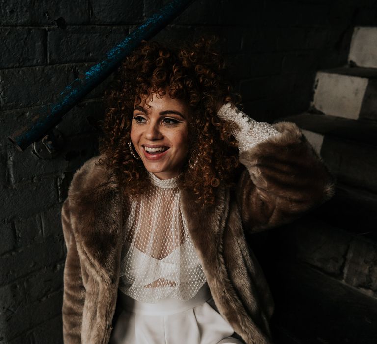 Bride with naturally curly hair in a bridal jumpsuit and brown faux fur coat 