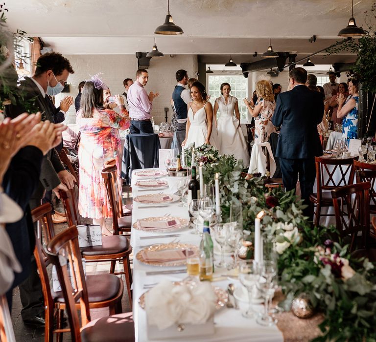 Bride in white cami wedding dress and bride in white lace top capped sleeve wedding dress walk into wedding reception room full of guests stood around tables with foliage table runners and white candles at The West Mill Derby