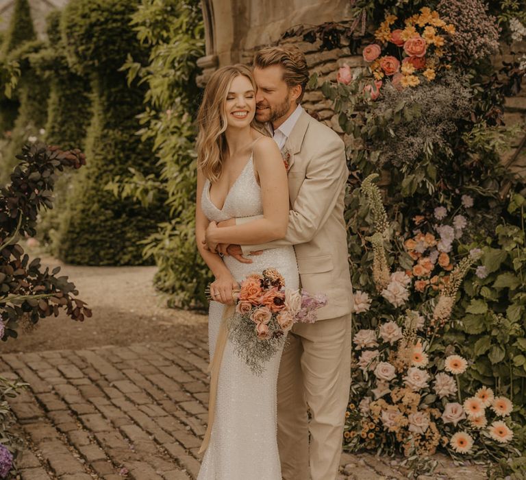 Groom in a beige suit embracing his bride in a sequin Made With Love wedding dress by the castle arch at Euridge Manor 