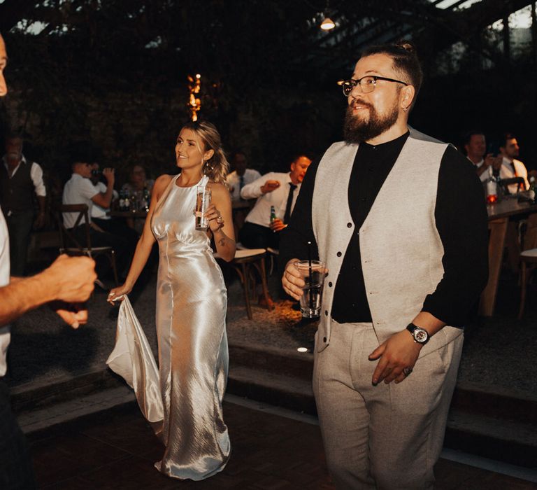 Bride with curled ponytail holds drink and train of her silver satin dress for wedding evening party at the glasshouse at Anran Devon