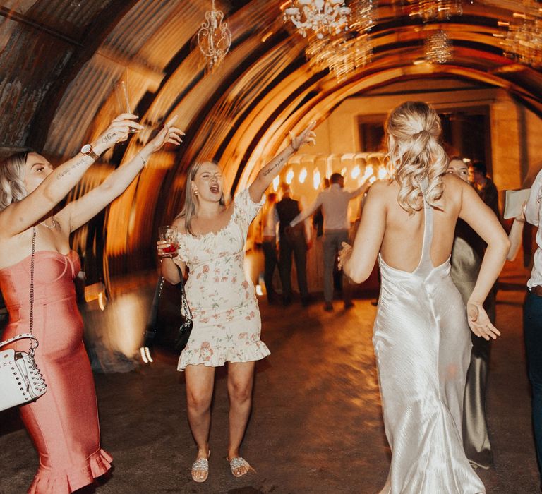 Bride in racer back silver satin dress and curled blonde ponytail dances with wedding guests in the hut lit by chandeliers at Anran Devon