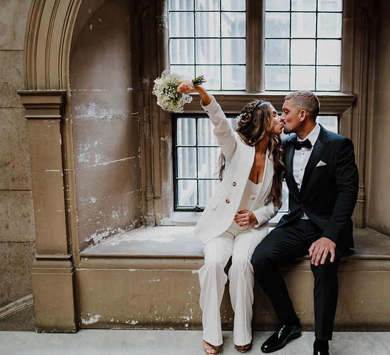 Groom in a black tuxedo kissing his bride in a white suit holding a white rose and gypsophila bouquet 