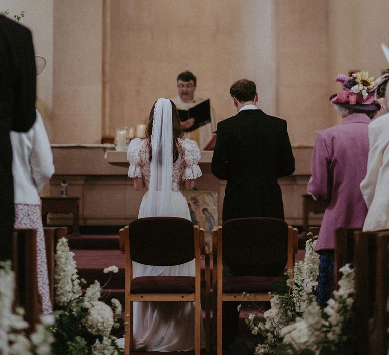 Bride in white lace puffed sleeve Daalarna wedding dress and bespoke veil stands next to groom in black morning coat at the end of the aisle during church wedding ceremony