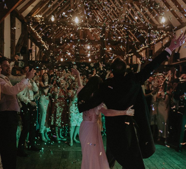 Bride in white lace puffed sleeve wedding dress hugs groom in black morning coat as confetti falls on them and wedding guests in beamed room at Loseley Park in Surrey