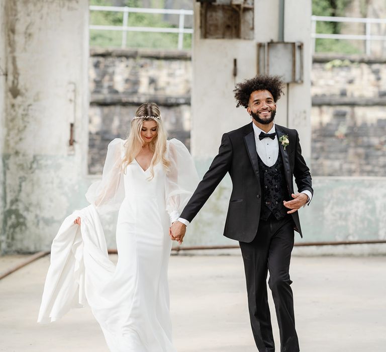 A couple walk hand in hand laughing. The bride wears a white dress and the groom wears black tie.