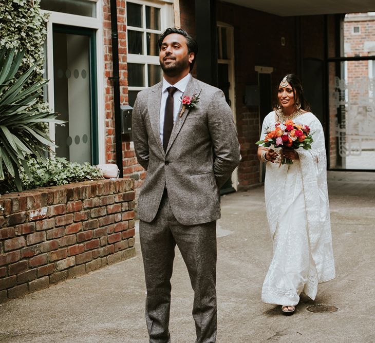 A first look for Indian British wedding. The groom wears a grey suit and the bride wears a white sari and carries a colourful bouquet.