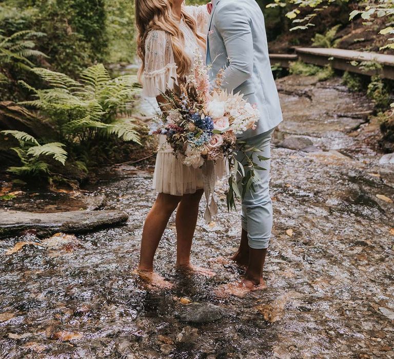 Bride & stand barefoot in waterfall after wedding ceremony outdoors 