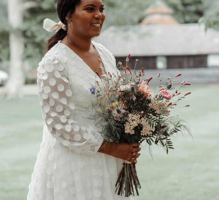 Bride in long mesh sleeved spotted Vagabond wedding dress and bridal hair bow holding mixed fresh and dried bouquet before Wasing Park wedding ceremony