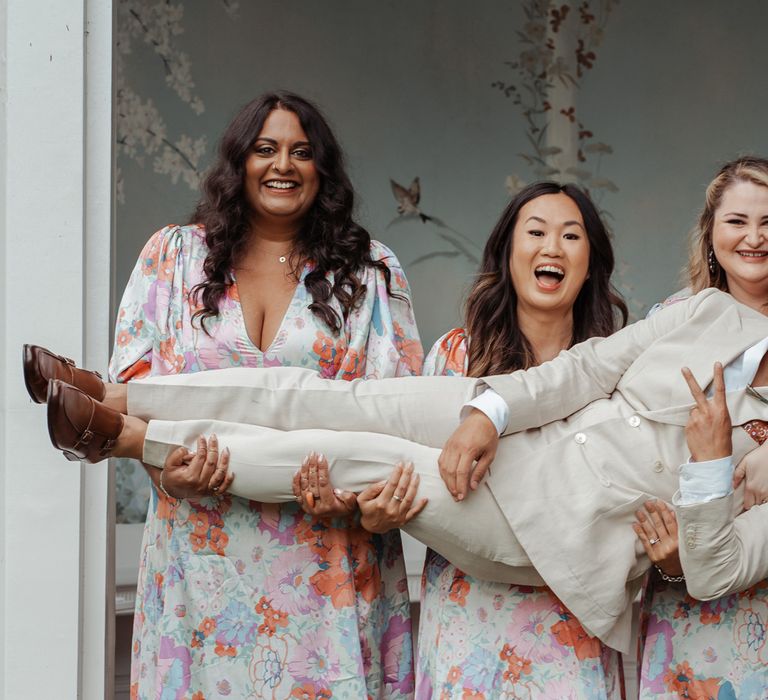 Three bridesmaids in V neck floral bridesmaid dresses hold up groom in double breasted linen suit and brown shoes wearing sunglasses outside Victorian summerhouse at Wasing Park wedding