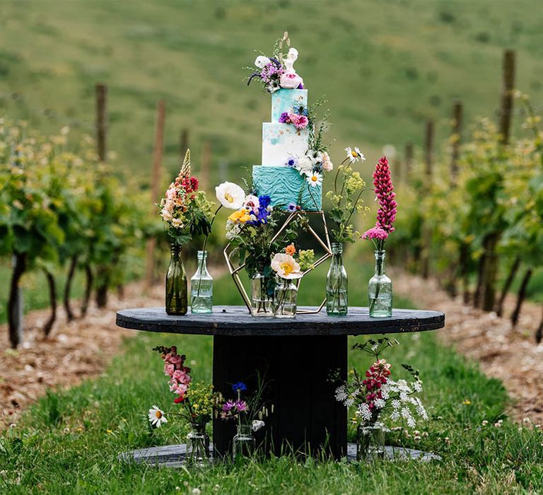 Three-tier turquoise and white buttercream wedding cake wresting on a cotton wheel surrounded by colourful wildflowers at Little Wold Vineyard