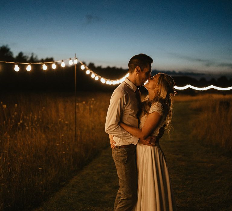 Bride in lace top wedding dress with train hugs groom in white shirt with grey trousers stand in field with festoon lighting during late summer wedding at Wellington Wood Norfolk