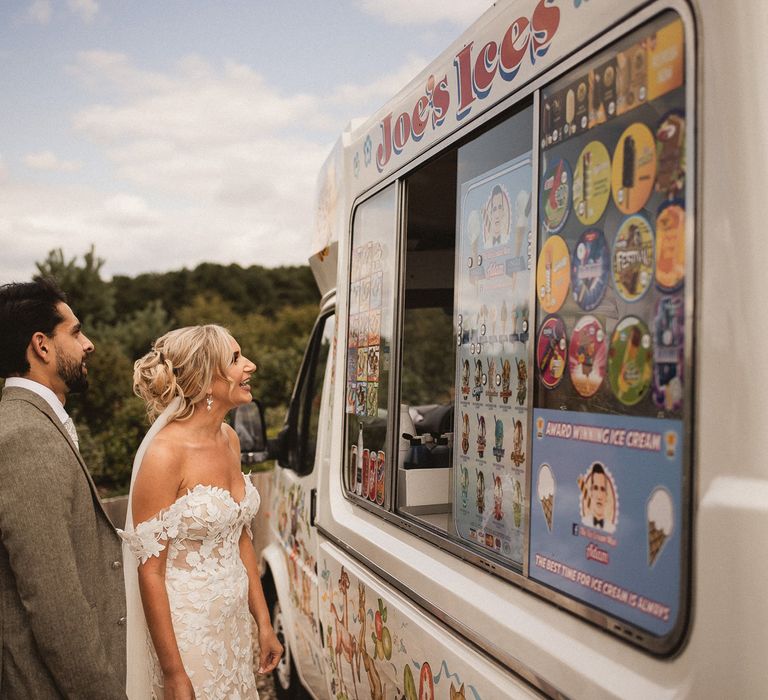 Bride in off the shoulder Enzoani wedding dress and groom in grey suit stand at Ice cream van during tipi wedding at Inkersall Grange Farm